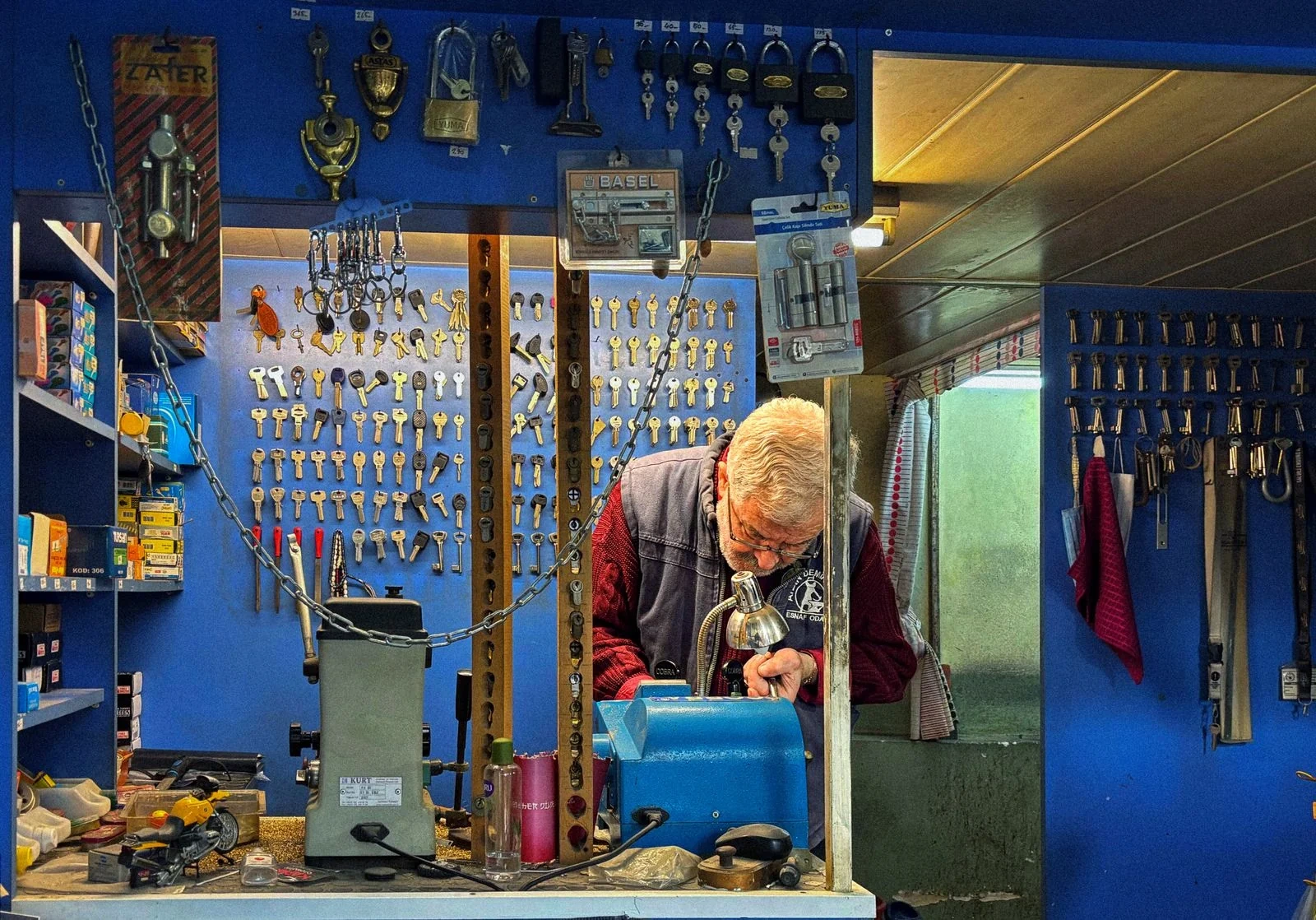 Person leaning over a blue bench machine in a small shop with keys, padlocks and door hardware displayed on blue walls.