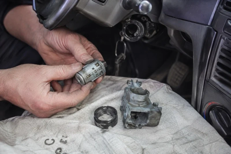 Hands holding a removed ignition cylinder beside a car steering column, with the housing and small parts on a cloth.