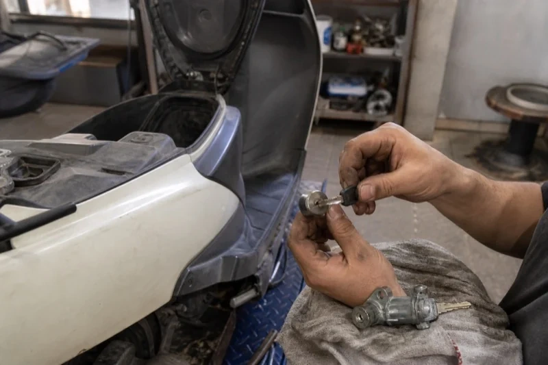 Hands holding a key and removed lock cylinder beside a scooter with its seat raised, with another lock assembly on a cloth.